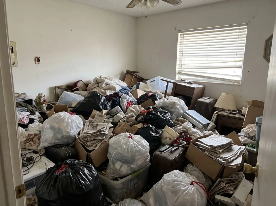 Illustration: a spare room in a Florida house filled waist-high with bags, newspapers, boxes, clothing and small furniture so the floor cannot be seen, daylight through the window blinds.