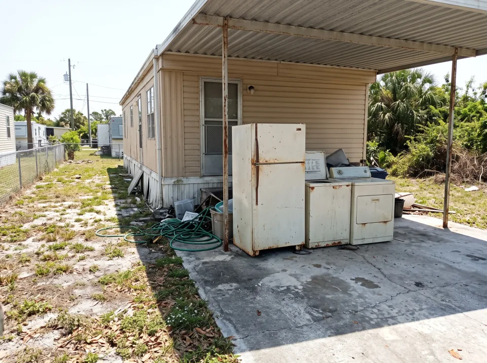Illustration: an old refrigerator, a washer and a dryer pushed together in the carport at a sand beige mobile home in a Belle Glade park with a carport and small yard, clear midday sun