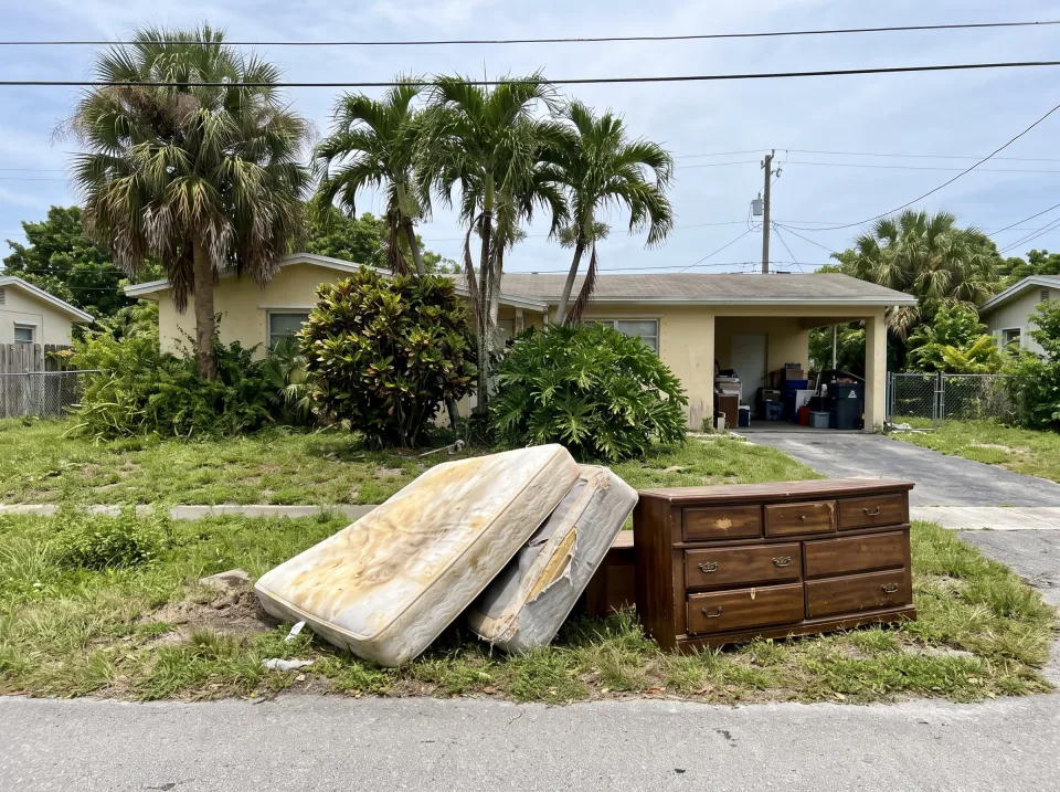 Illustration: a two-car garage in a South Florida ranch house packed wall to wall with cardboard boxes, an old treadmill, a broken recliner, paint cans, bikes and bags of clothes, roll-up door open to bright daylight.