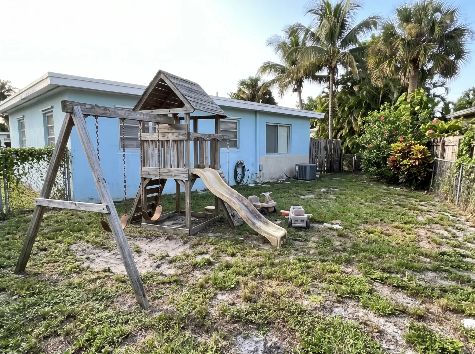 Illustration: a sun-faded wooden swing set and playhouse in the backyard at a sky blue single-story Coral Terrace concrete block home with a flat white roof and palm trees, bright morning light