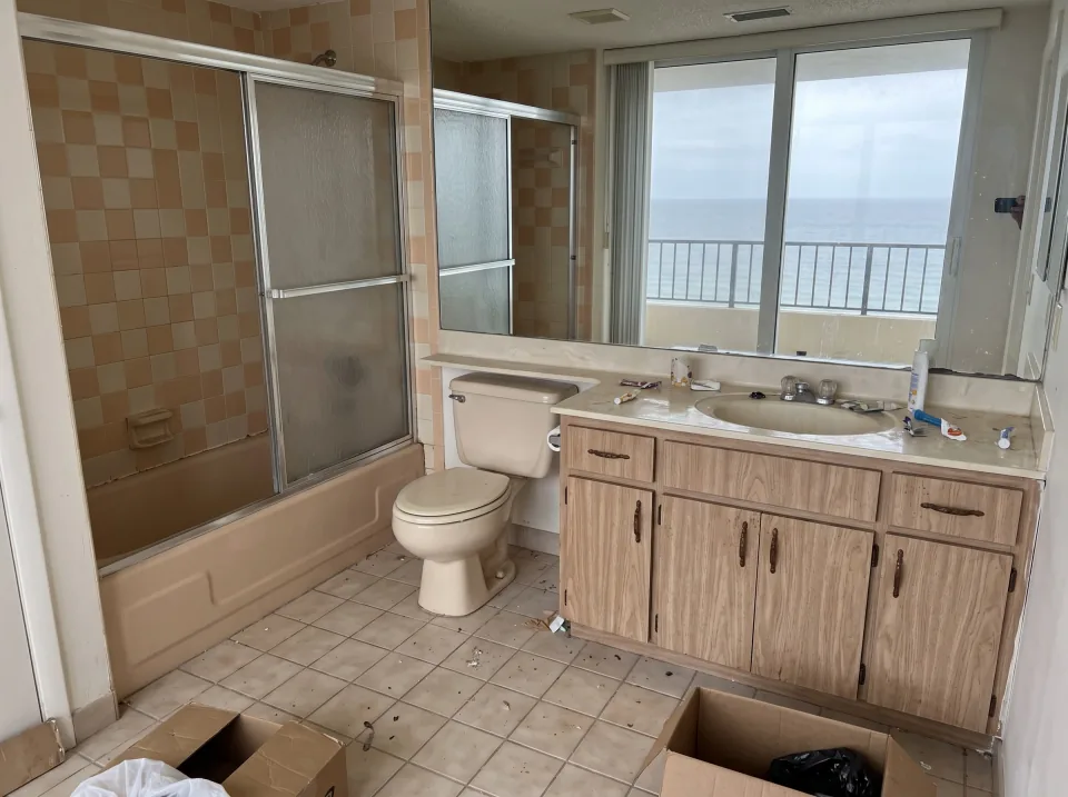 Illustration: a dated tiled bathroom with an old tub, toilet and vanity inside a Golden Beach beachside condo unit with ocean view through sliding glass doors, soft overcast light