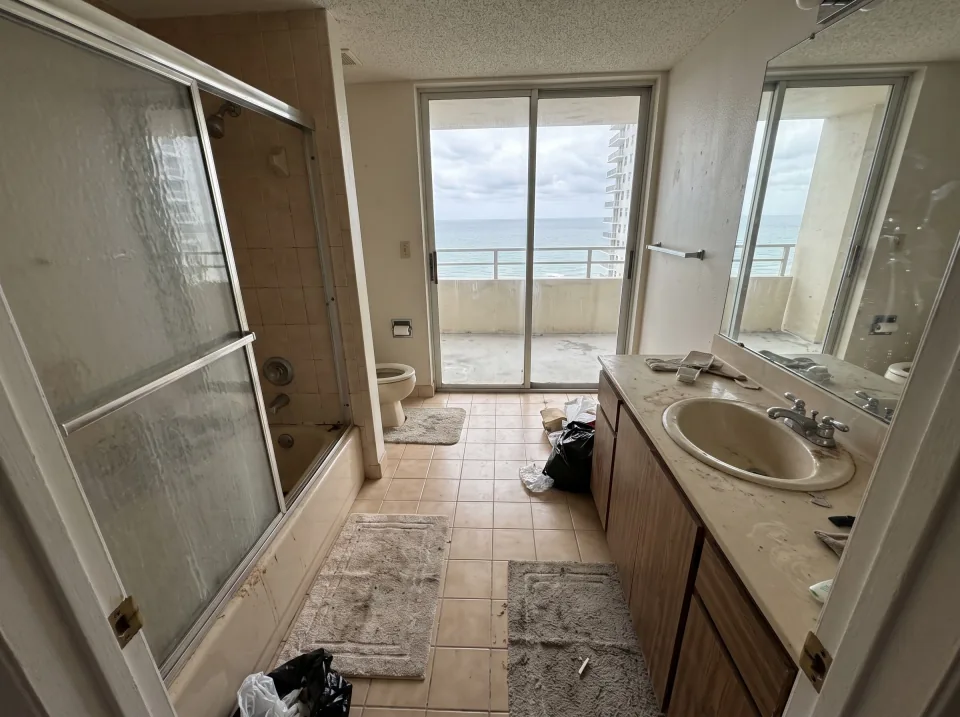 Illustration: a dated tiled bathroom with an old tub, toilet and vanity inside a Hallandale Beach beachside condo unit with ocean view through sliding glass doors, soft overcast light