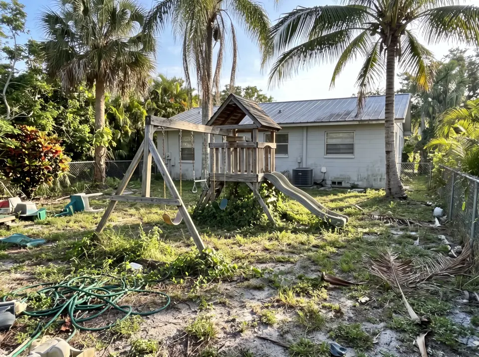 Illustration: a sun-faded wooden swing set and playhouse in the backyard at a light gray single-story Haverhill concrete block home with a standing-seam metal roof and palm trees, bright morning light