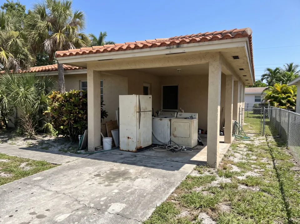 Illustration: an old refrigerator, a washer and a dryer pushed together in the carport at a sand beige single-story Hialeah Gardens concrete block home with a terracotta tile roof and palm trees, clear midday sun