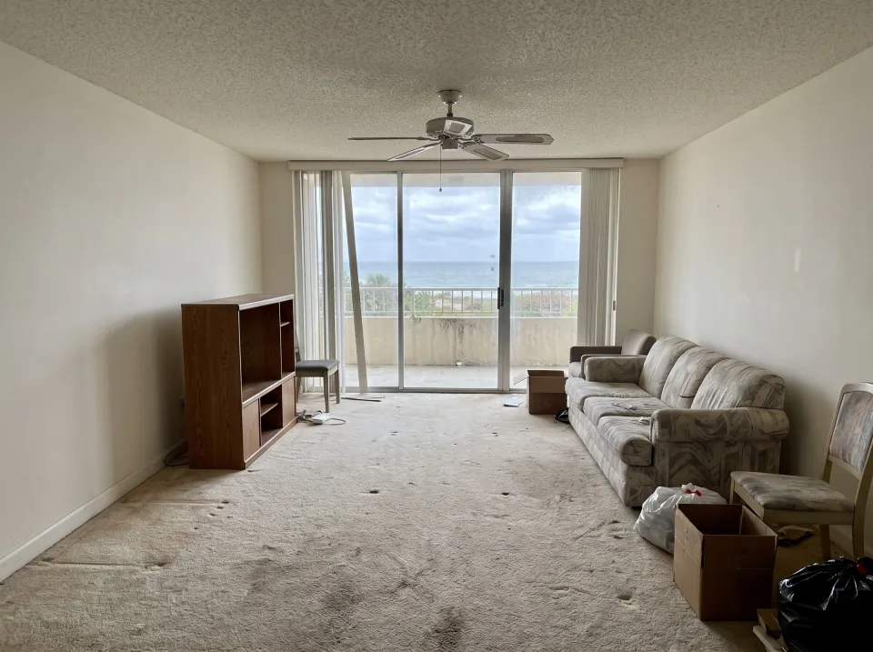 Illustration: a living room with old wall-to-wall carpet and a few leftover pieces of furniture inside a Highland Beach beachside condo unit with ocean view through sliding glass doors, soft overcast light