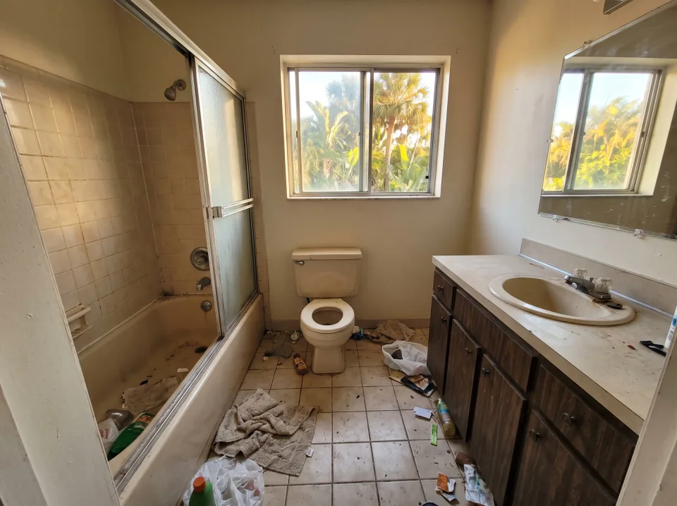 Illustration: a dated tiled bathroom with an old tub, toilet and vanity at a light gray single-story Weston concrete block home with a flat white roof and palm trees, warm late-afternoon light