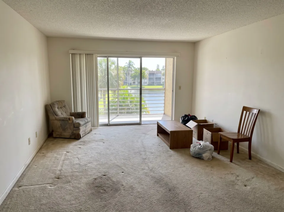 Illustration: a living room with old wall-to-wall carpet and a few leftover pieces of furniture inside a Lauderdale Lakes garden-style condo unit with a screened balcony looking out on palm trees and a lake, clear midday sun
