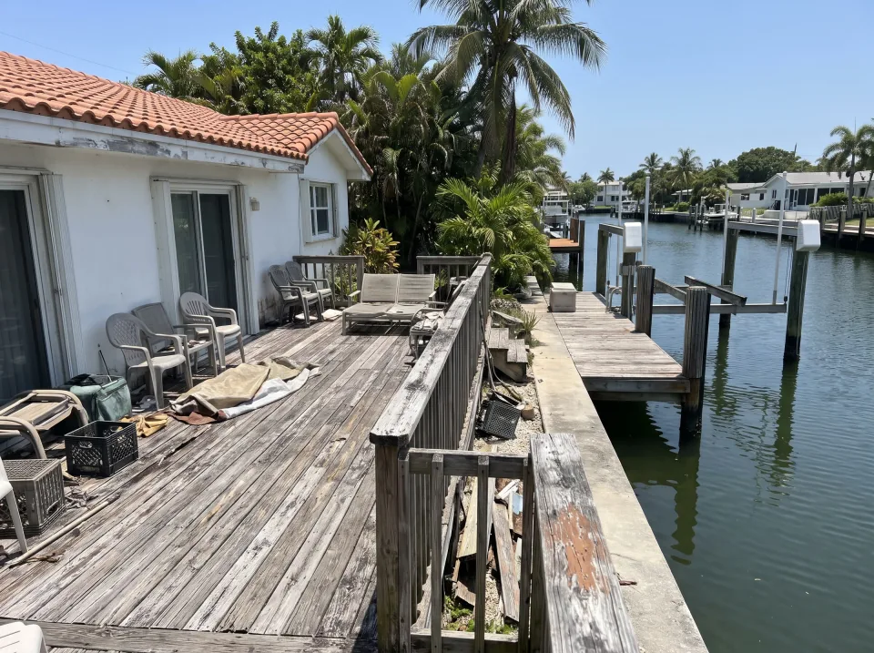 Illustration: an old weathered wooden deck with railings behind the house at a white Lighthouse Point canal-front home with a boat dock and calm water, terracotta tile roof, clear midday sun