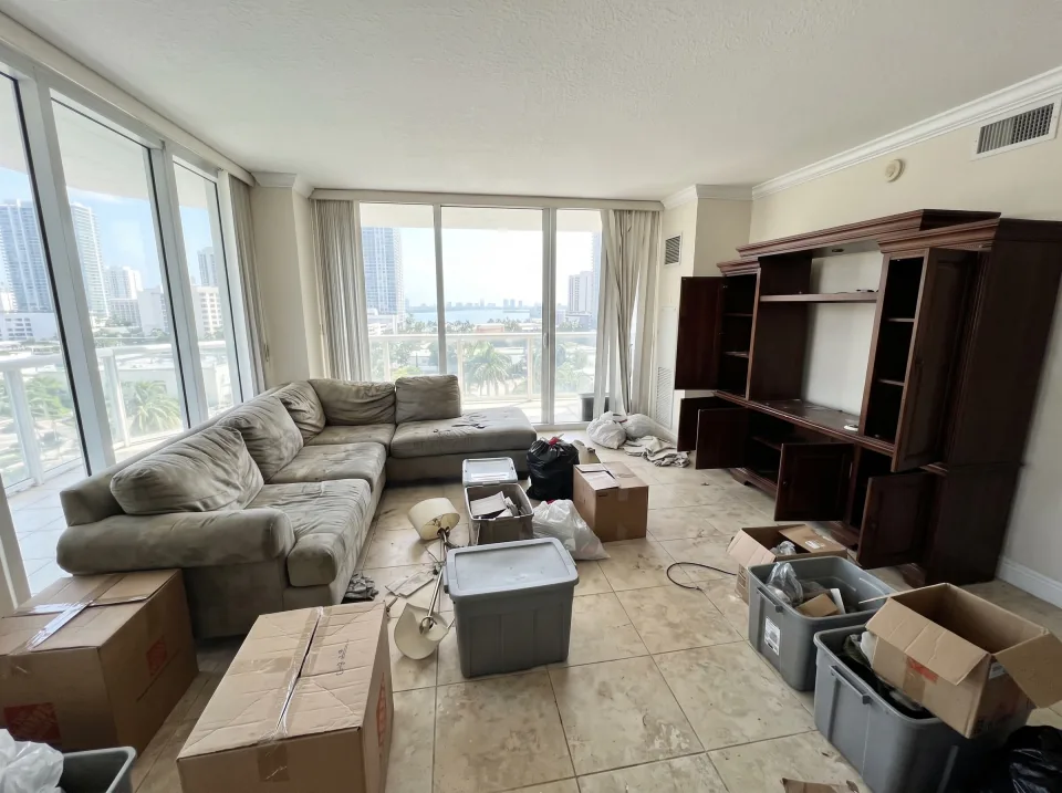 Illustration: a dated living room in a 1970s Florida house with terrazzo floors: a worn sectional sofa, a heavy wooden entertainment center, a broken recliner and a stained area rug.