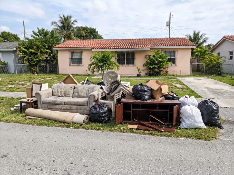 Illustration: a large curbside pile of old furniture, a mattress, carpet rolls and black trash bags on the grass swale of a South Florida street.