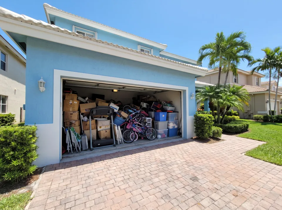 Illustration: a garage packed with boxes, an old treadmill, bikes and storage bins at a sky blue two-story home in a Miami Lakes gated community with manicured landscaping, flat white roof, clear midday sun
