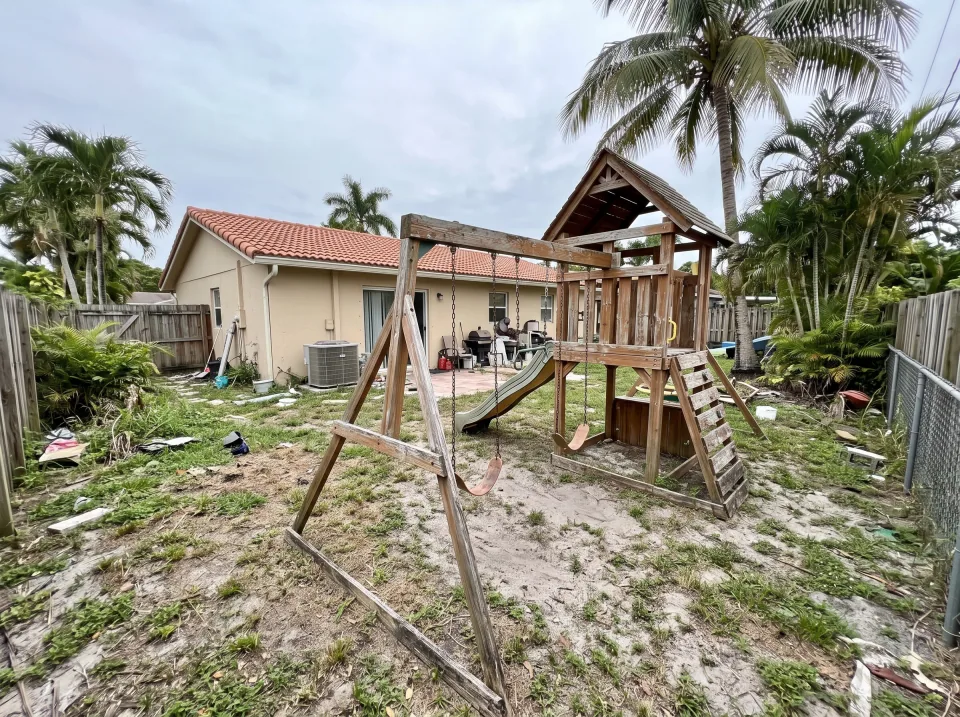 Illustration: a sun-faded wooden swing set and playhouse in the backyard at a sand beige single-story North Lauderdale concrete block home with a terracotta tile roof and palm trees, soft overcast light