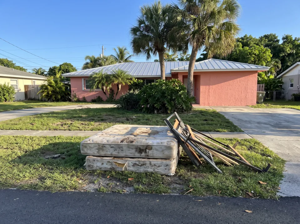 Illustration: an old stained mattress, a box spring and a broken bed frame left on the grass swale at a coral pink single-story Palmetto Bay concrete block home with a standing-seam metal roof and palm trees, bright morning light