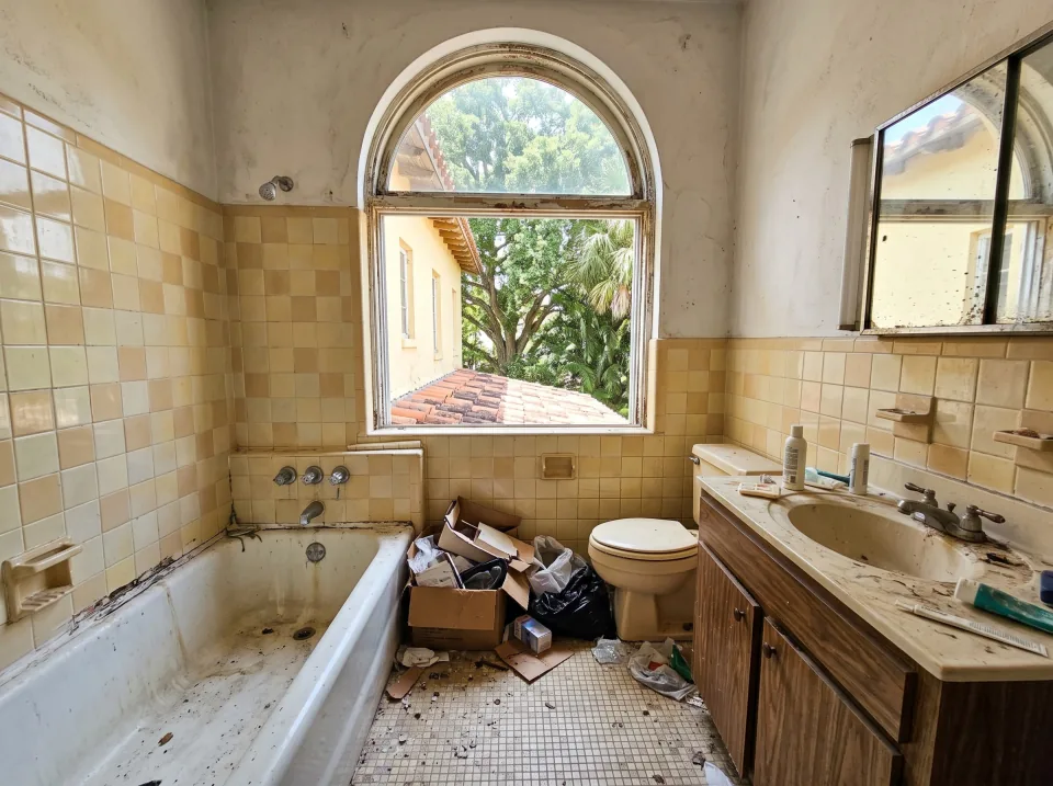 Illustration: a dated tiled bathroom with an old tub, toilet and vanity at a pale yellow historic South Miami home with arched windows, terracotta tile roof and big shade trees, clear midday sun