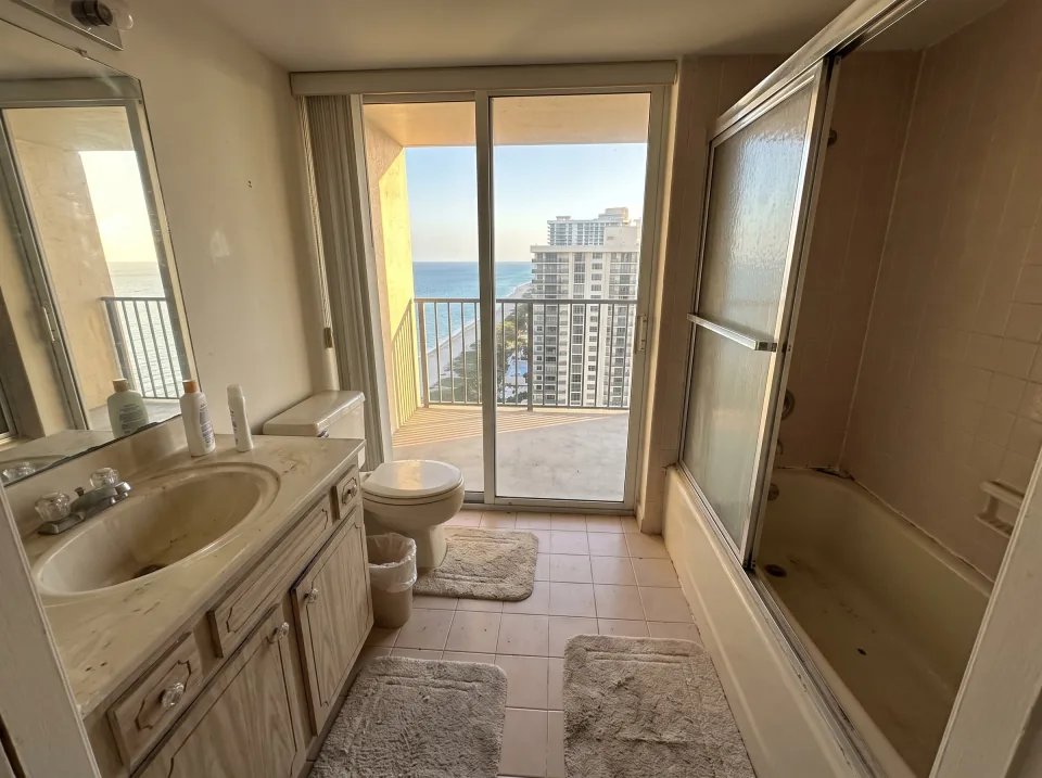 Illustration: a dated tiled bathroom with an old tub, toilet and vanity inside a Sunny Isles Beach beachside condo unit with ocean view through sliding glass doors, warm late-afternoon light