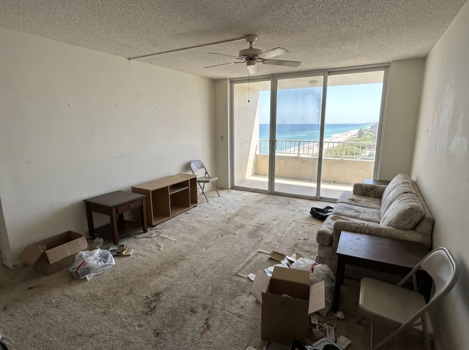 Illustration: a living room with old wall-to-wall carpet and a few leftover pieces of furniture inside a Surfside beachside condo unit with ocean view through sliding glass doors, clear midday sun