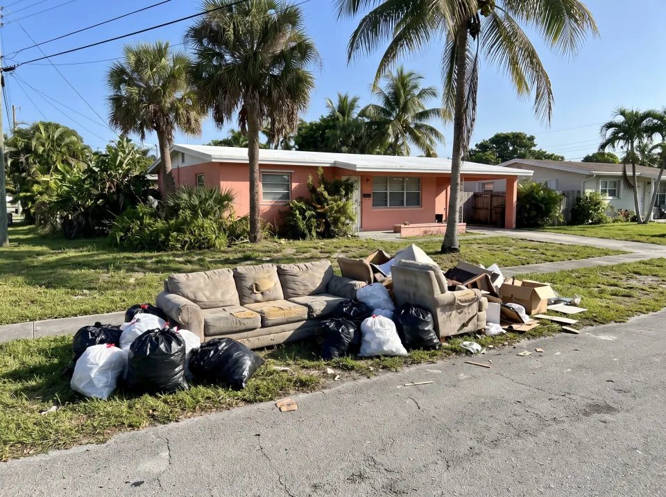 Illustration: a worn sectional sofa, an armchair and bagged junk piled at the curb at a coral pink single-story West Park concrete block home with a flat white roof and palm trees, bright morning light