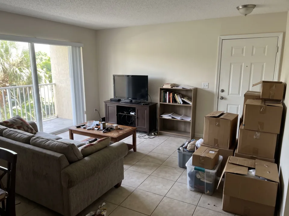 Illustration: a furnished South Florida apartment living room with a sofa, coffee table, TV stand, bookshelf and moving boxes stacked by the door.